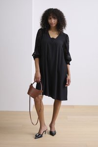 Vestido negro con mangas de encaje, escote en V, corte holgado; combinado con un bolso marrón y zuecos negros de tacón con acentos decorativos.
