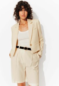 Blazer beige à bouton unique, coupe ajustée, porté sur un débardeur blanc. Associé à un short beige taille haute plissé et une ceinture noire.