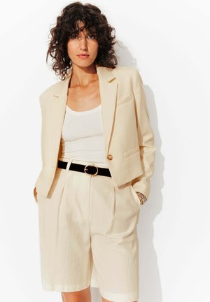Blazer beige à bouton unique, coupe ajustée, porté sur un débardeur blanc. Associé à un short beige taille haute plissé et une ceinture noire.