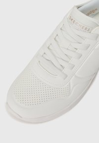 White sneaker made of smooth synthetic material, featuring a perforated toe box, flat laces, and a logo label on the tongue.