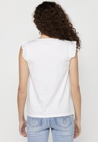 Top in cotone bianco con maniche in pizzo arricciato, scollatura rotonda e vestibilità comoda. Indossato con jeans in denim blu, mostrando un semplice design sul retro.
