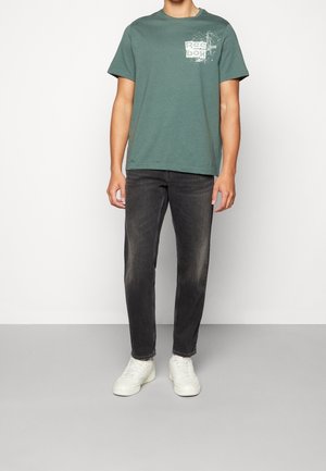 Uomo che indossa una maglietta Reebok verde con grafica, jeans neri sbiaditi e sneakers bianche, in piedi davanti a uno sfondo chiaro e semplice.