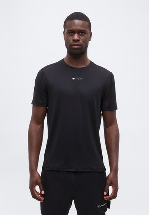 Mand iført sort Champion t-shirt og shorts, stående front mod en ensfarvet hvid baggrund.
