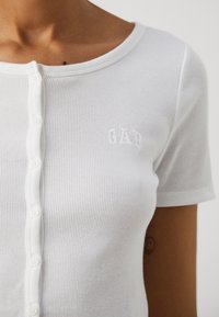 Vit ribbad t-shirt med utkärning, korta ärmar och framknäppning; har en liten, vit broderad logotyp på vänster sida.