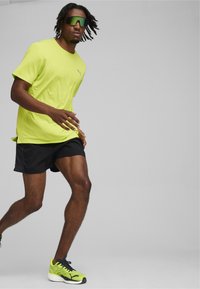 Camiseta atlética de color amarillo neón de Puma con mangas cortas, combinada con pantalones cortos negros. Lleva gafas de sol con tintado verde y calzado a juego con detalles negros.
