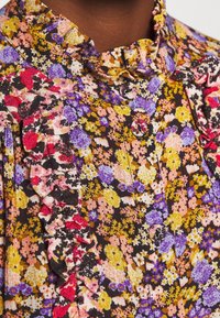 Bluse med blomsterprint, der har en sort baggrund med farverige blomster i pink, lilla og gul; volangkrave og knapdetalje.