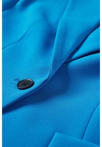 Tissu bleu vif avec un seul bouton noir et des coutures visibles, montrant une partie d'un blazer ou d'un vêtement sur mesure à l'avant.
