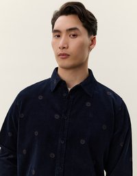 Camicia in corduroy blu navy con complessi ricami circolari, chiusura con bottoni, colletto classico e maniche lunghe. Texture e dettagli sottili.