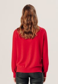 Roter gestrickter Pullover mit langen Ärmeln und rundem Ausschnitt. Weicher Stoff, leicht lockere Passform und gerippter Saum. Getragen mit schwarzen Lederhosen.