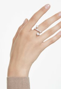 Eine Hand, die drei Ringe trägt: einen Perlenring, einen Diamantring und einen Ring mit einer Reihe kleiner Edelsteine. Die Hand ist vor einem schlichten Hintergrund positioniert.