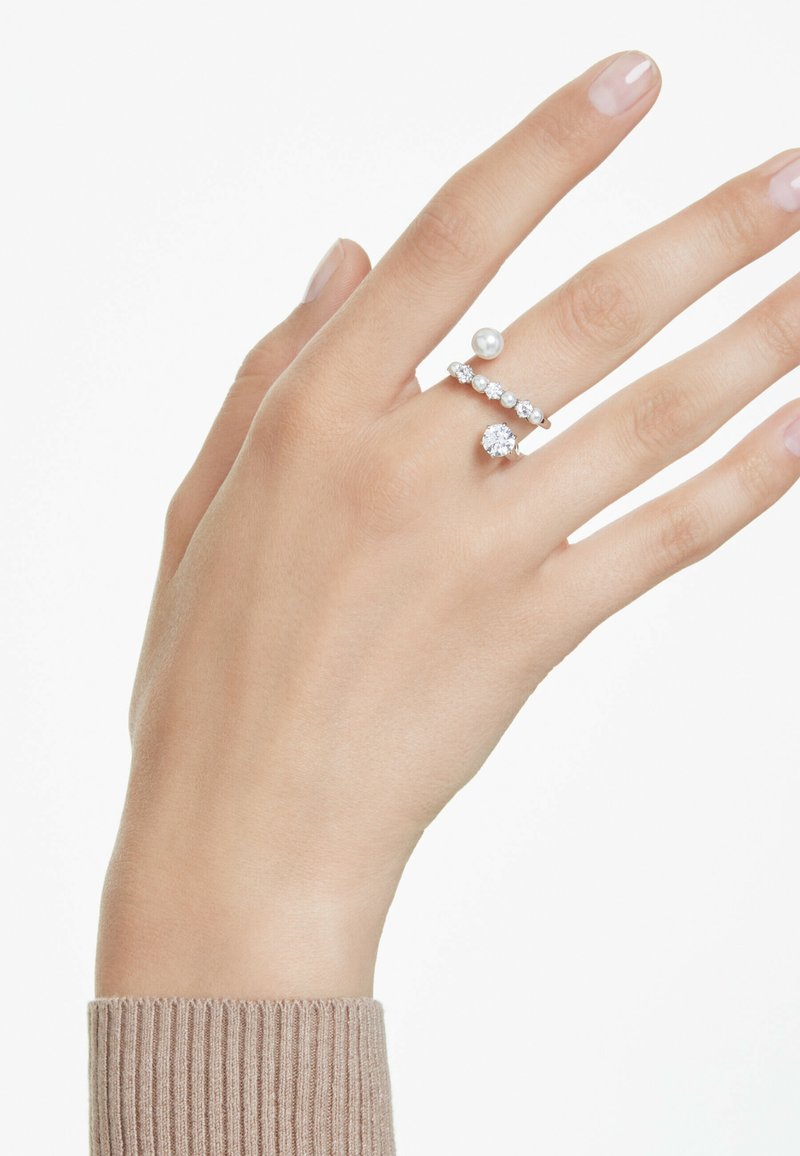Eine Hand, die drei Ringe trägt: einen Perlenring, einen Diamantring und einen Ring mit einer Reihe kleiner Edelsteine. Die Hand ist vor einem schlichten Hintergrund positioniert.