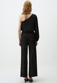 Combinaison noire avec une épaule et une manche longue, dotée d'une texture côtelée et de pantalons à jambes larges.