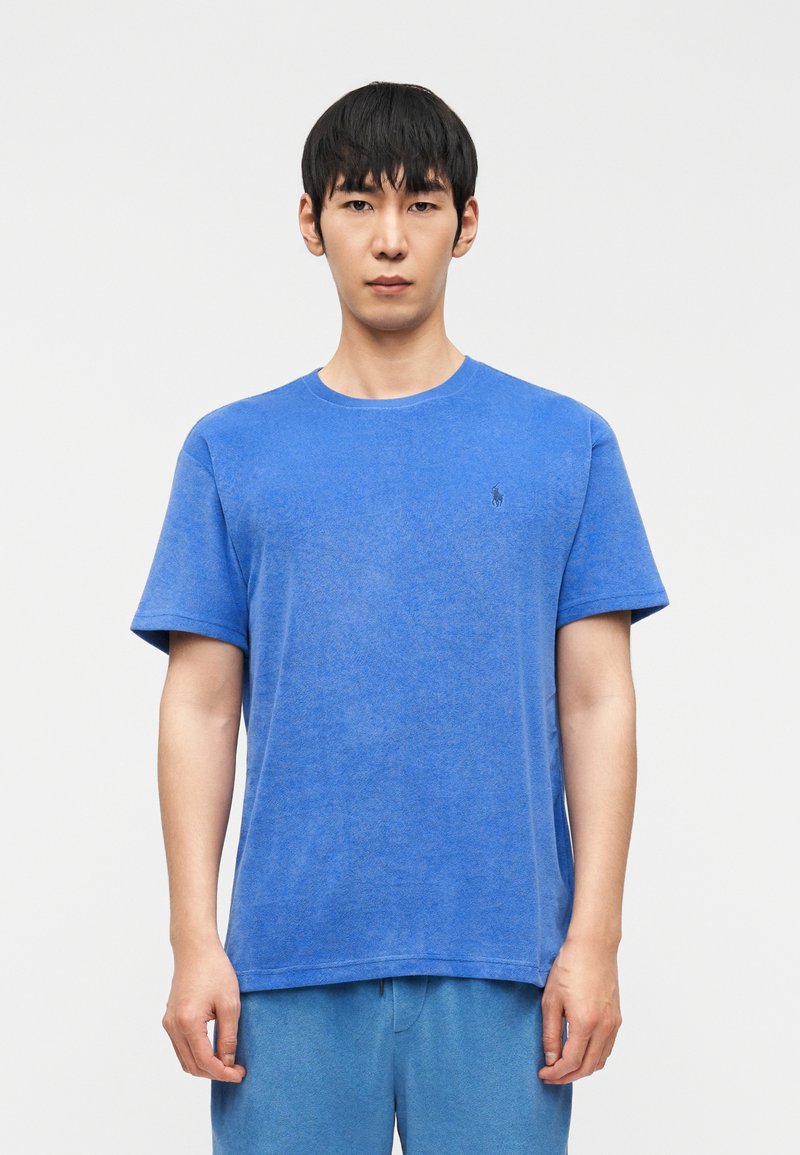 Νεαρός άνδρας που στέκεται στραμμένος προς τα εμπρός, φορώντας ένα απλό μπλε κοντομάνικο πουκάμισο και ταιριαστό μπλε παντελόνι, με φόντο ένα λευκό τοίχο.