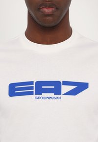 Balta medvilnės marškinėliai su ryškiai mėlynu tekstu "EA7" ir "EMPORIO ARMANI." Apvalaus kaklo dizainas, lygus paviršius, be matomų raštų ar akcentų.