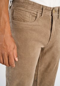 Brązowe spodnie z korduroju o teksturowej powierzchni, z przednią kieszenią, paskami na pasek oraz metalowym guzikiem w pasie.