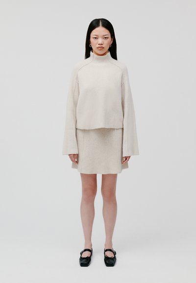 Maglione a coste beige con collo alto, abbinato a una gonna a coste coordinata. Il modello indossa scarpe nere con punta quadrata. Focus sulla texture e sui sovrapposizioni.