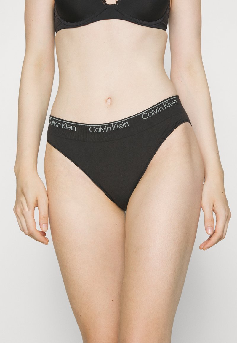 Tanga in cotone nero con un morbido cinturino elastico che presenta un logo grigio "Calvin Klein" ripetuto. Texture liscia, design minimalista.