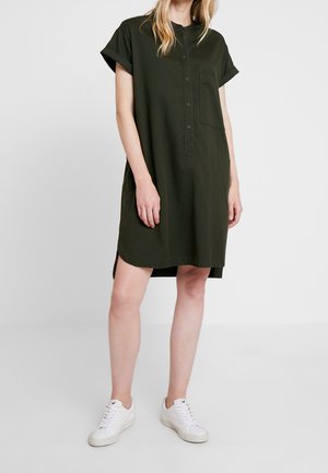 Femme portant une robe boutonnée à manches courtes de couleur vert foncé avec une poche avant et des baskets blanches, debout devant un fond uni.