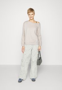 Hellgrauer, übergroßer Pullover, beigefarbene, weite Cargo-Hose mit Taschen, blaue Schuhe und eine kleine graue Handtasche. Glatte Stofftextur.