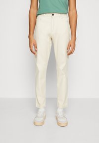 Osoba v off-white rovně střižených kalhotách, bílých teniskách s béžovými podrážkami a zeleném topu zastrčeném do kalhot, stojící před jednobarevným pozadím.