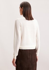 Sweter dzianinowy w kolorze off-white z długimi rękawami i prążkowanymi mankietami, charakteryzujący się klasycznym wzornictwem i miękką teksturą. Noszony z brązową zamszową spódnicą.