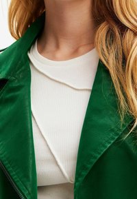 Veste en similicuir vert à large col portée par-dessus un haut blanc côtelé avec un col festonné et une texture douce.