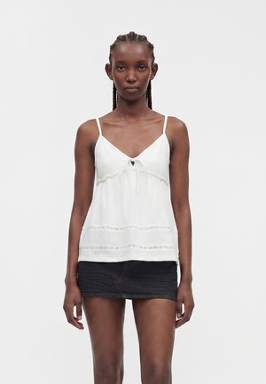 Femme aux cheveux tressés portant un haut blanc sans manches avec des détails en dentelle et une mini-jupe en jean noire, debout devant un fond uni.