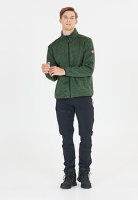 Giovane uomo in piedi, indossando una giacca verde con zip, pantaloni tattici neri e scarponi da trekking neri, contro uno sfondo bianco semplice.