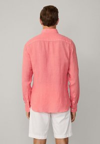 Camicia a maniche lunghe in lino corallo chiaro, con colletto classico, polsini con bottone e orlo curvo, indossata con pantaloni corti bianchi.