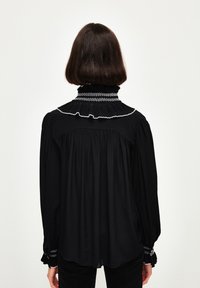 Schwarze Bluse mit hohem Rüschkragen, elastischen Bündchen und weißen Zickzacknähten. Fließender Stoff mit lockerer Passform.