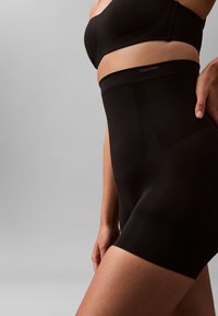 Frau trägt hoch taillierte schwarze Calvin Klein Shaping-Shorts und passenden schwarzen BH vor einem schlichten grauen Hintergrund.