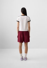 A person stands facing away, wearing a white shirt with black trim and burgundy shorts, paired with light socks and purple shoes.