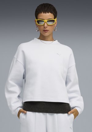 Korthvid sweatshirt med lange ærmer, blød tekstur, der har et subtilt logo og er lagdelt over en mørk skjorte. Kompletteret med gule solbriller.