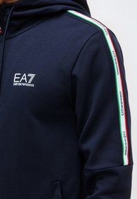 Sudadera con capucha azul marino con el logo EA7 Emporio Armani en el pecho y cinta con franjas verde, blanco y rojo con branding a lo largo del hombro y la manga.