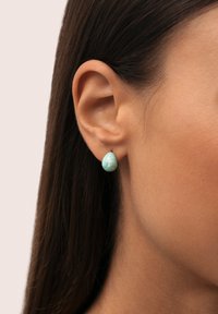 Pendiente en forma de lágrima en turquesa con una textura veteada, montado en un pequeño poste de metal, llevado en la oreja de un modelo. El fondo es claro.