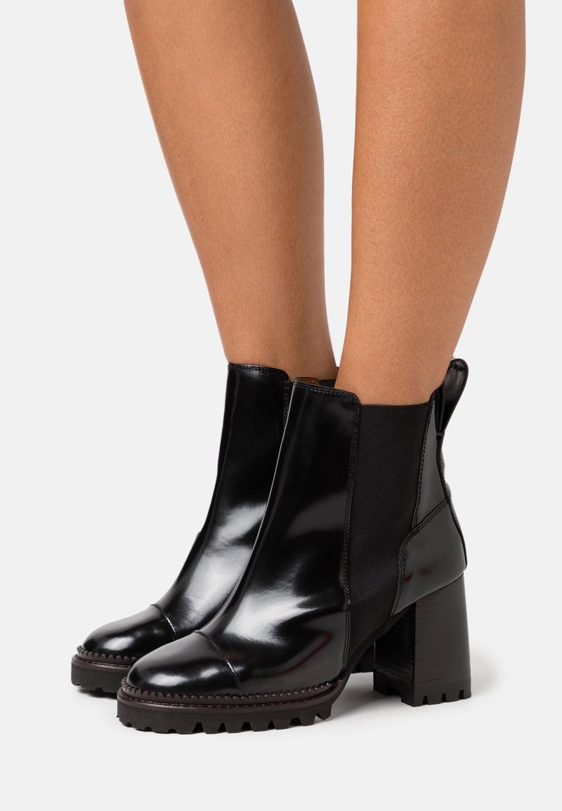 Bottes courtes en cuir noir brillant avec panneaux latéraux élastiques, talons blocs épais et semelles robustes portées sur des jambes nues sur un fond uni.