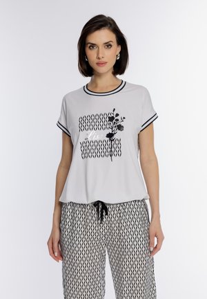 Kobieta ubrana w biały krótki top z czarnym kwiatowym wzorem i tekstem, w połączeniu z luźnymi spodniami w czarno-biały wzór, stojąca na białym tle.