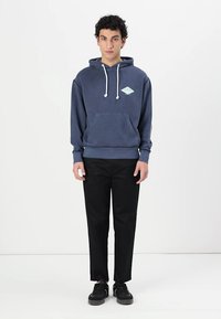 Felpa in cotone blu navy con lacci bianchi, tasca frontale e badge logo azzurro. Abbinata a pantaloni neri e sneakers nere.