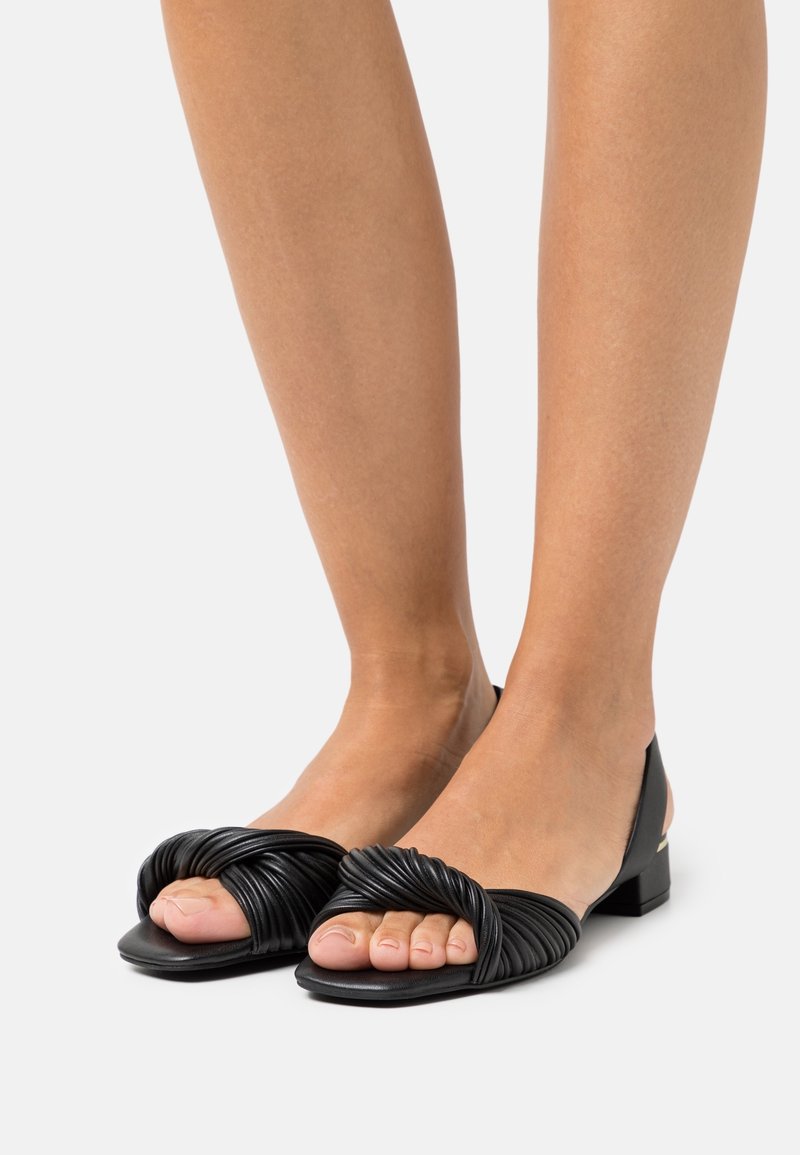 Pieds portant des sandales plates noires à bout ouvert avec un design de sangle torsadée et un détail de talon fermé sur un fond clair uni.