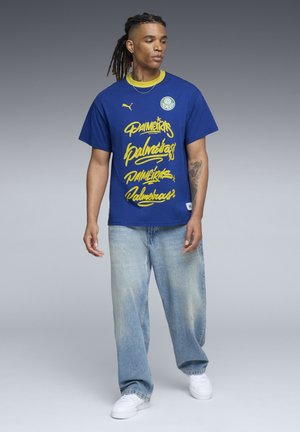 Blå bomulds t-shirt med gul graffiti-lignende tekst med "Palmeiras," rundt logo på venstre bryst, parret med lyseblå jeans og hvide sko.