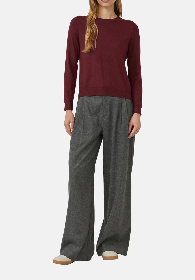 Woman wearing a burgundy long-sleeve top, gray wide-leg trousers, and beige shoes standing against a white background.