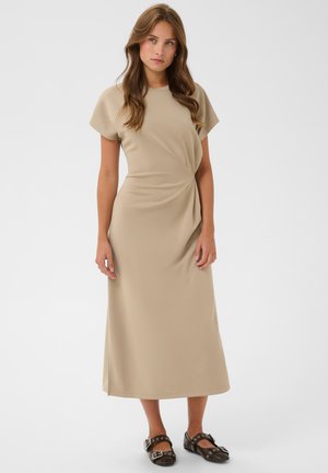 Femme aux longs cheveux bruns portant une robe midi beige à manches courtes et des chaussures noires à boucle, debout sur un fond blanc.