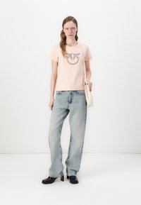Lys rosa t-skjorte med et dekorativt sølvlogo, kombinert med lyseblå, vide jeans og svarte hælboots. Nøytral bakgrunn.