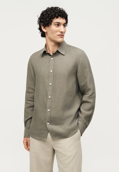 Άνδρας με σγουρά μαλλιά που φοράει μακρυμάνικο ελιάς πουκάμισο με κουμπιά και ανοιχτό μπεζ παντελόνι, κοιτάει προς τα πλάγια με τα χέρια στις τσέπες.