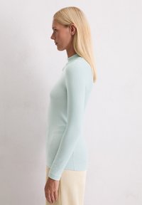 Helles blaues, langärmliges, geripptes Oberteil mit hohem Kragen; enganliegendes Design. Das Model posiert seitlich vor einem schlichten, strukturierten Hintergrund.