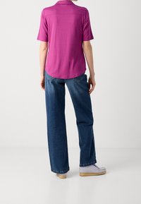 Paarse polo met korte mouwen en een ronde onderrand, gecombineerd met losse blauwe spijkerbroek en lichtgrijze schoenen, gezien vanuit de achterkant.