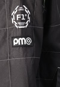 Black racing suit fabric features quilted texture, white stitching, and two patches: an embroidered F1 logo and a rectangular pm@ label.
