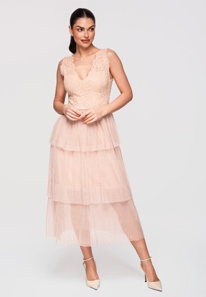 Mujer con cabello oscuro que lleva un vestido midi en tonos rosa rubor sin mangas de encaje y tul en capas y tacones nude con correa en el tobillo, de pie con las manos entrelazadas.