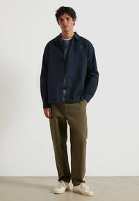 Giacca button-up blu navy con colletto, indossata sopra un maglione azzurro chiaro. I pantaloni verde oliva e le sneakers crema completano l'outfit.