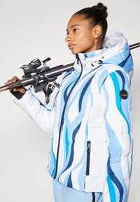 Blaue und weiße Daunenjacke mit Wellenmuster, Reißverschluss vorne und Kapuze. Hält ein Paar Skier mit Bindungen. Leicht und isoliert.